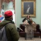 man dressed in pelts stands in capitol while a man with confederate flag looks on