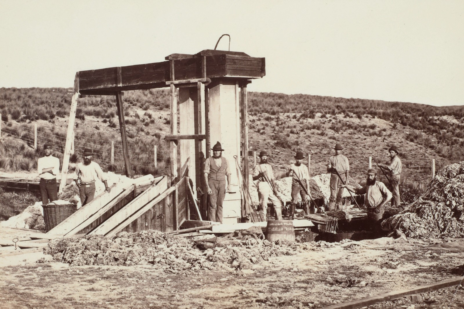 Eight men stand at an outdoor work station beside piles of wool.