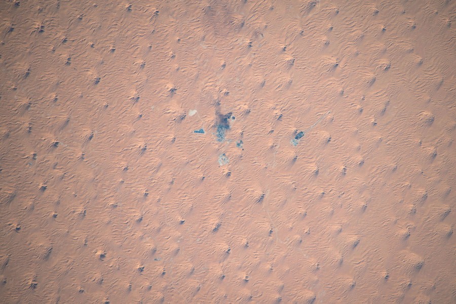 An orbital view of a desert dotted by dunes, with a dark blotch at center, where an industrial complex sits