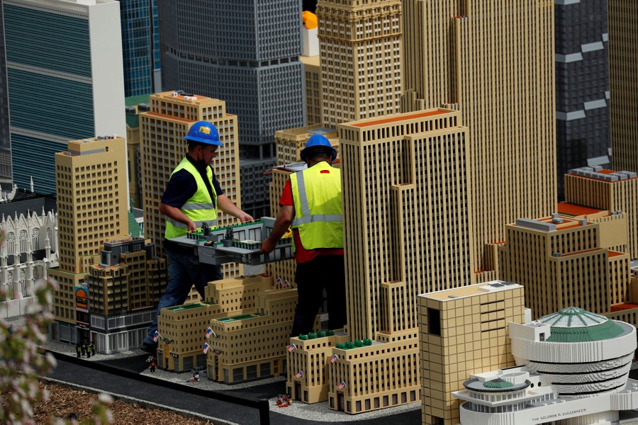 Workers carry a scale model of a structure among a cityscape of lego models.