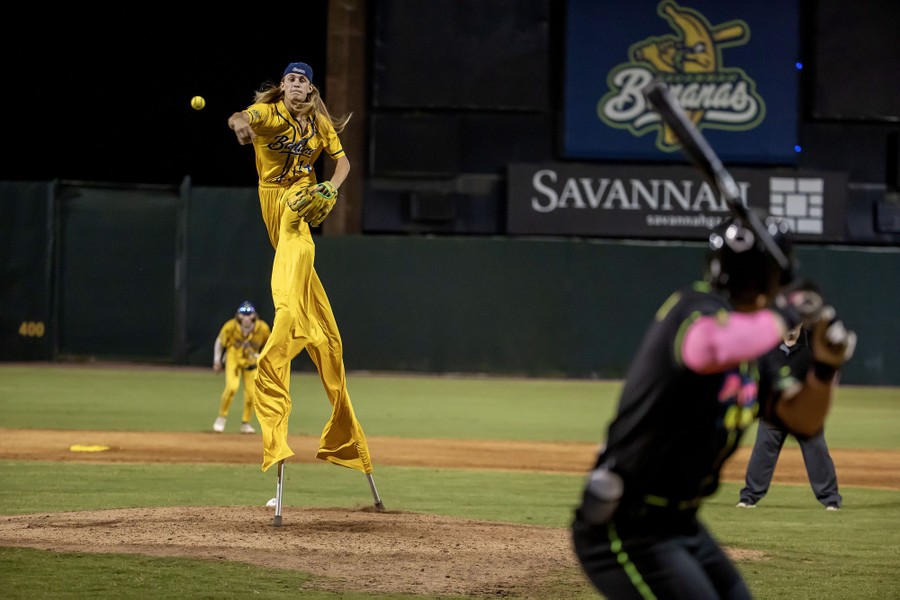 A baseball player wearing stilts pitches from a mound during a baseball game.