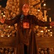 Ralph Fiennes as Dr. Kelson stands in front of an upside-down cross and a tall pile of skulls