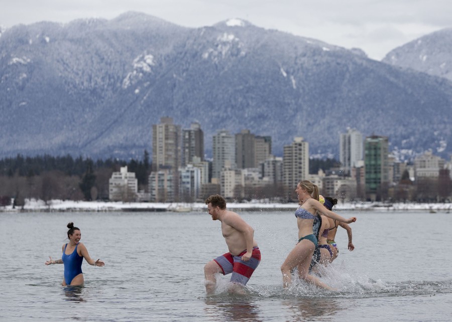 Several people run into a shallow bay, with a city and snowy mountains visible in the background.
