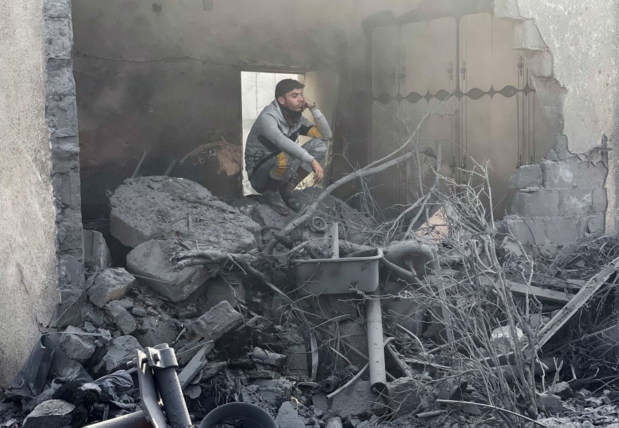 A person crouches atop a pile of rubble beside the smashed walls of a building.