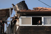 A color photograph of a man peering out the window of a house whose roof appears to have been destroyed by an explosion.