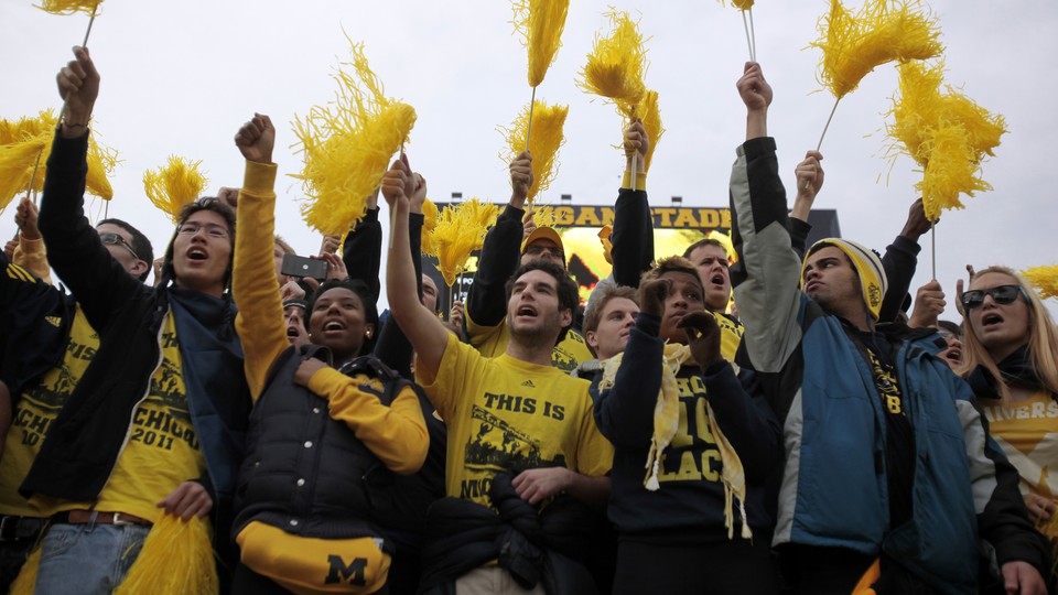 University of Michigan students hold yellow pom-poms and cheer in the stands of a football game. 