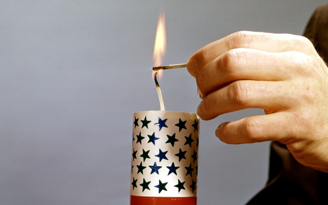A person's hands holding a red-white-and-blue firecracker while lighting the wick with a match