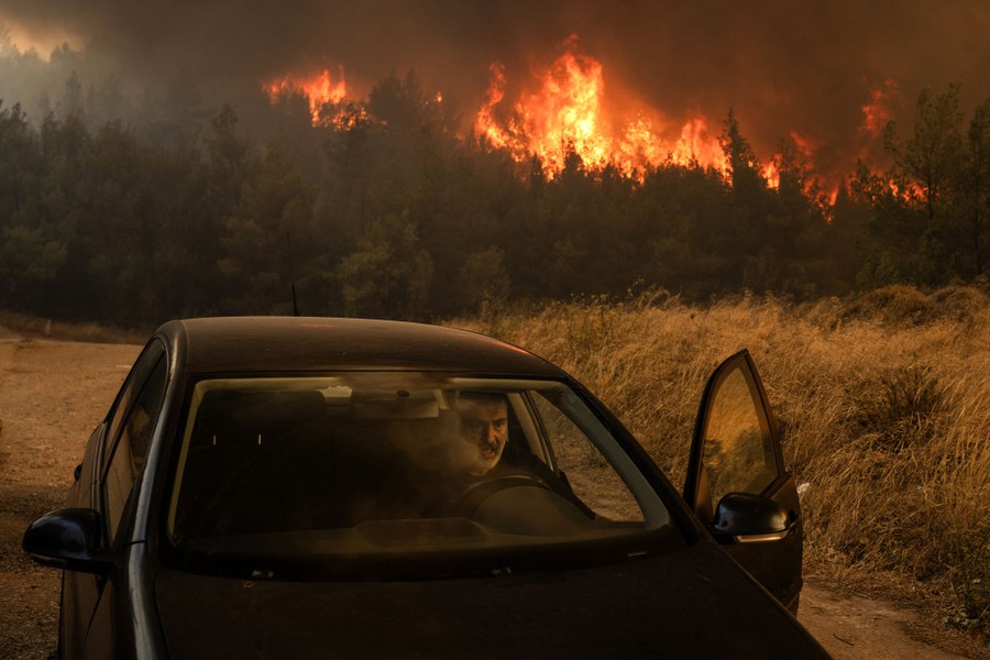A man in the driver's seat of a car, with a wildfire raging in the trees behind him