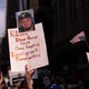 A hand holds up a sign that reads “Release Kilmar Abrego Garcia / Cease targeting Immigrant Communities” at a protest where others wave signs and upside-down American flags