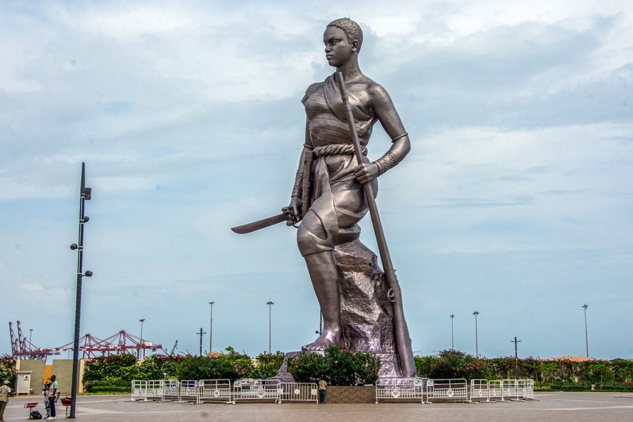 A tall metal statue depicts a woman holding a rifle and a sword.