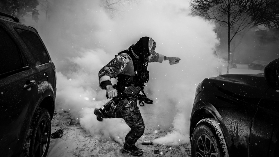 A black-and-white photo of an ICE agent between two cars and a cloud of tear gas in the background