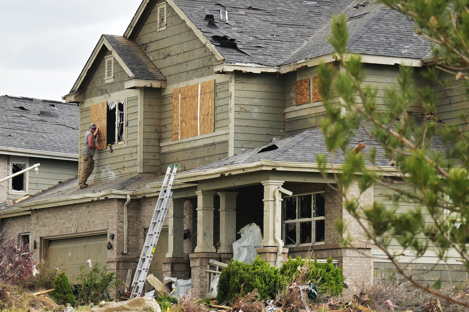 A worker stands on a roof, boarding up windows on a storm-damaged house.