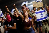 Protesters stands together in a street, holding up signs, Israeli flags, and their fists.