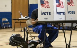 A child in a carrier sits next to their father as he votes. The father's face is covered by a partition that reads "VOTE" and is decorated with an American flag.