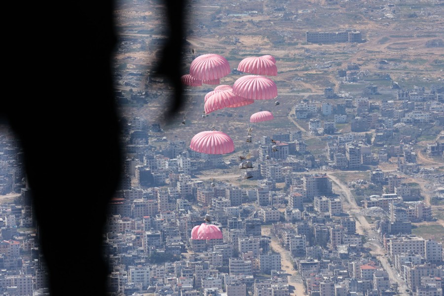 Crates of aid hanging from parachutes drop toward a city below, seen from the open back of a cargo aircraft.