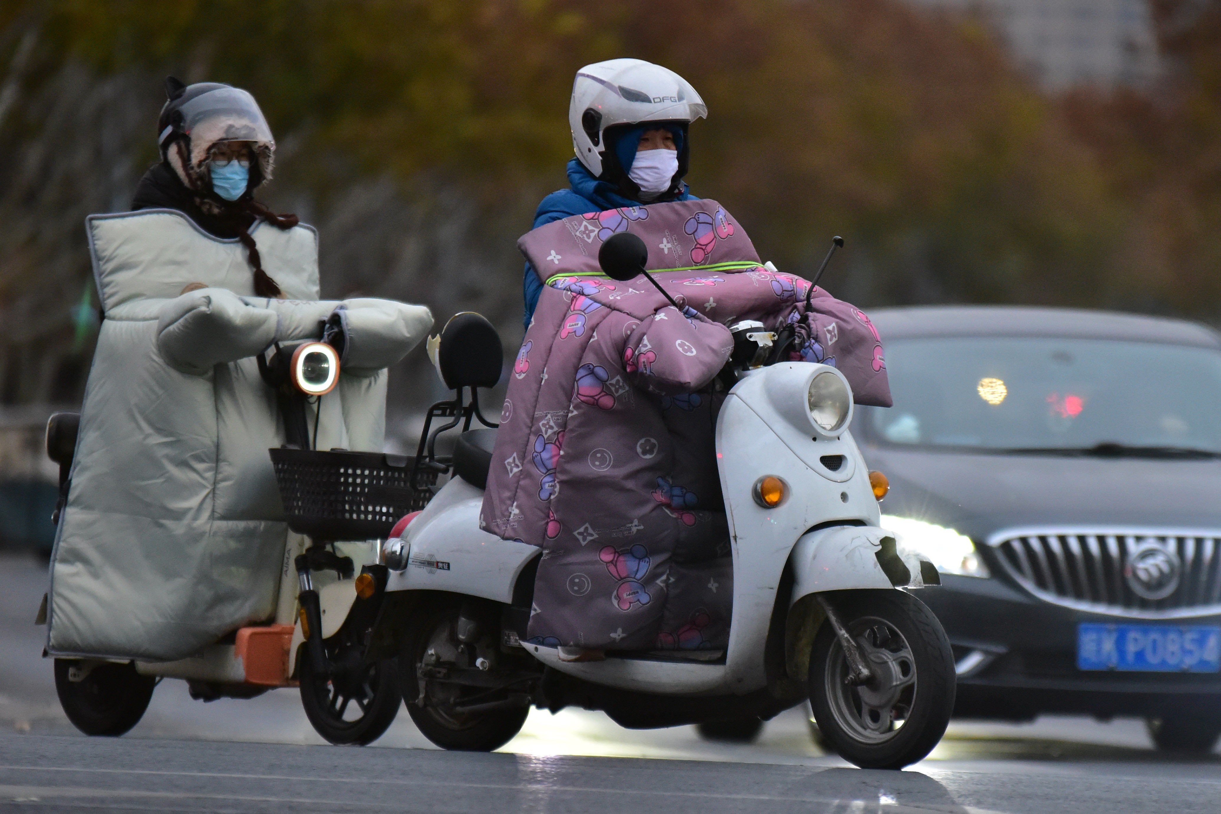 Two people ride on scooters, with custom-made blankets wrapped over their hands, handlebars, and the front of their body.