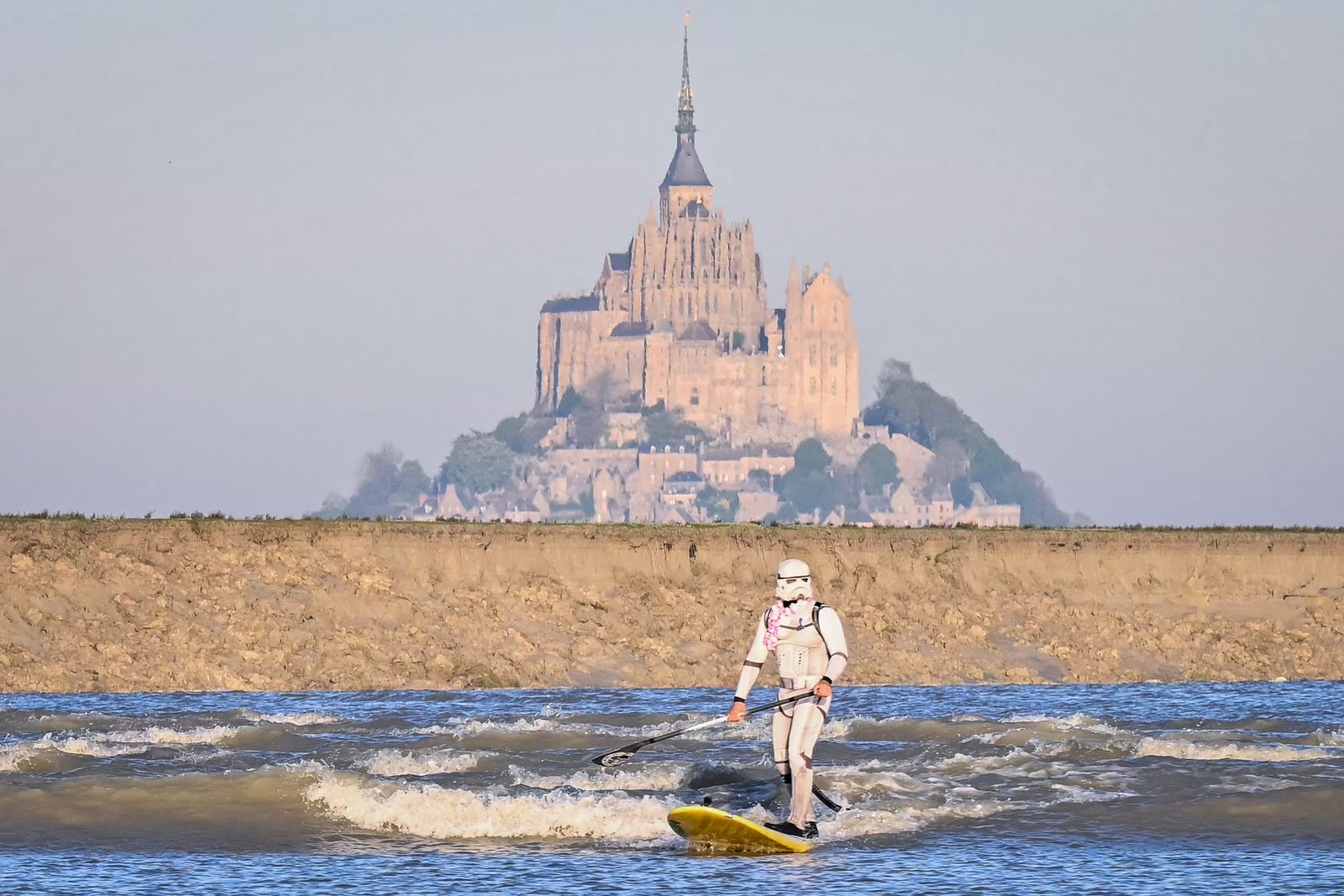 A surfer wearing a stormtrooper costume rides a wave in front of a distant historic building.