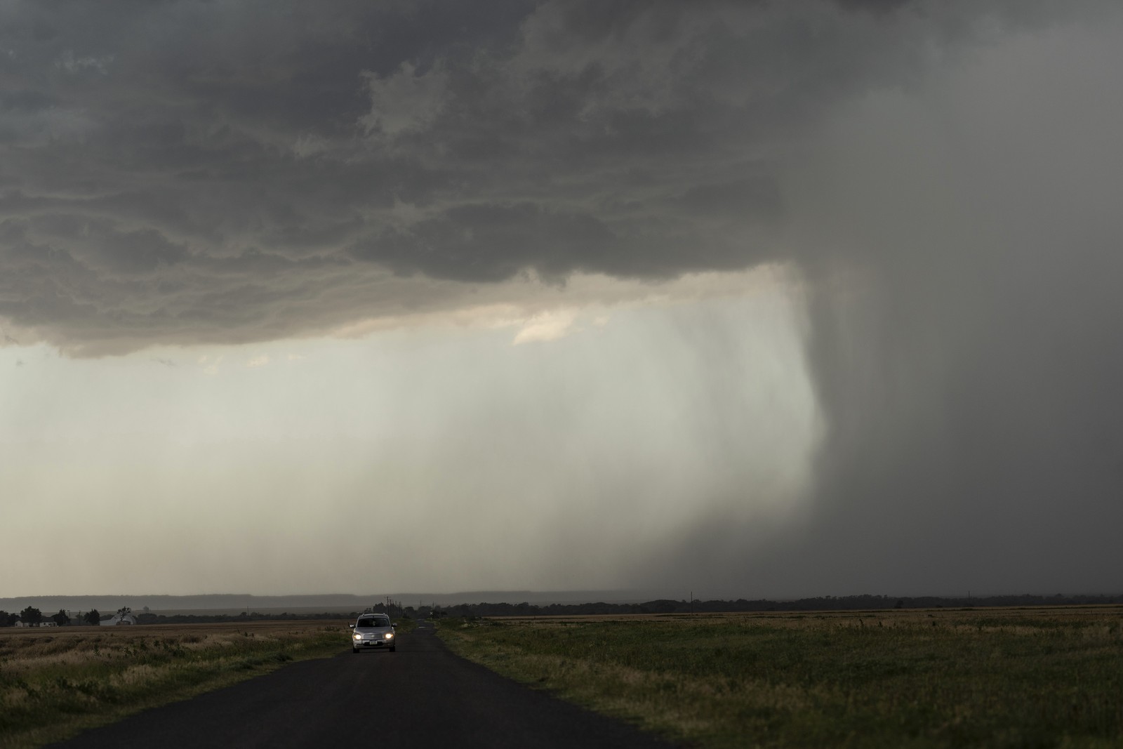 A car drives on a road near a visible downpour of raing beneath a dark storm cloud.
