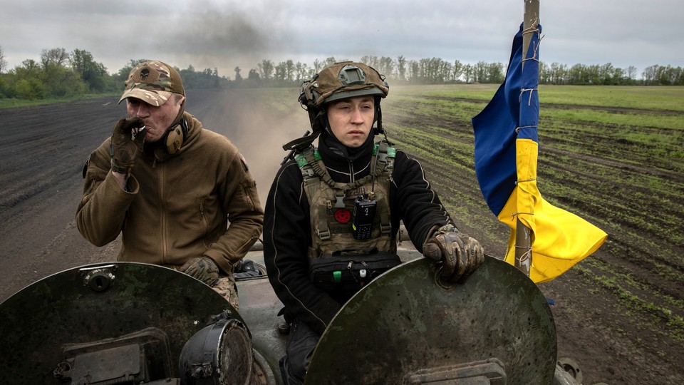 Two members of the Ukrainian 28th Mechanized Brigade ride in an armored vehicle towards hostile Russian forces. The one on the left is smoking a cigarette, the one on the right is scowling, and to their side is a blue and yellow flag.