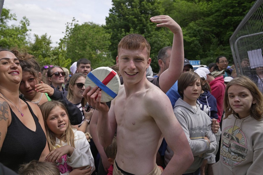 A man smiles while holding up a wheel of cheese wrapped in red and blue ribbons, with a crowd of onlookers behind him.