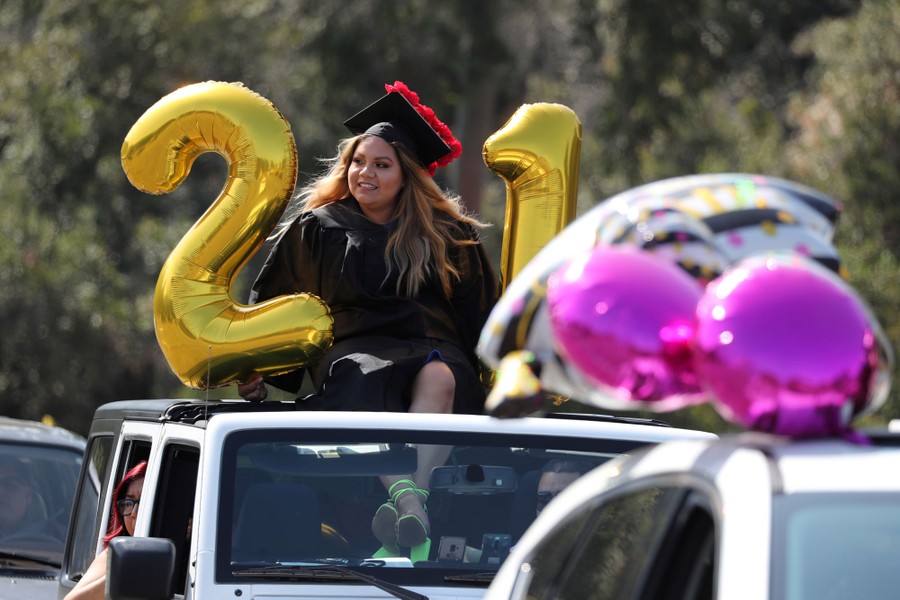 A graduating student rides atop a car, wearing a cap and gown, surrounded by balloons.