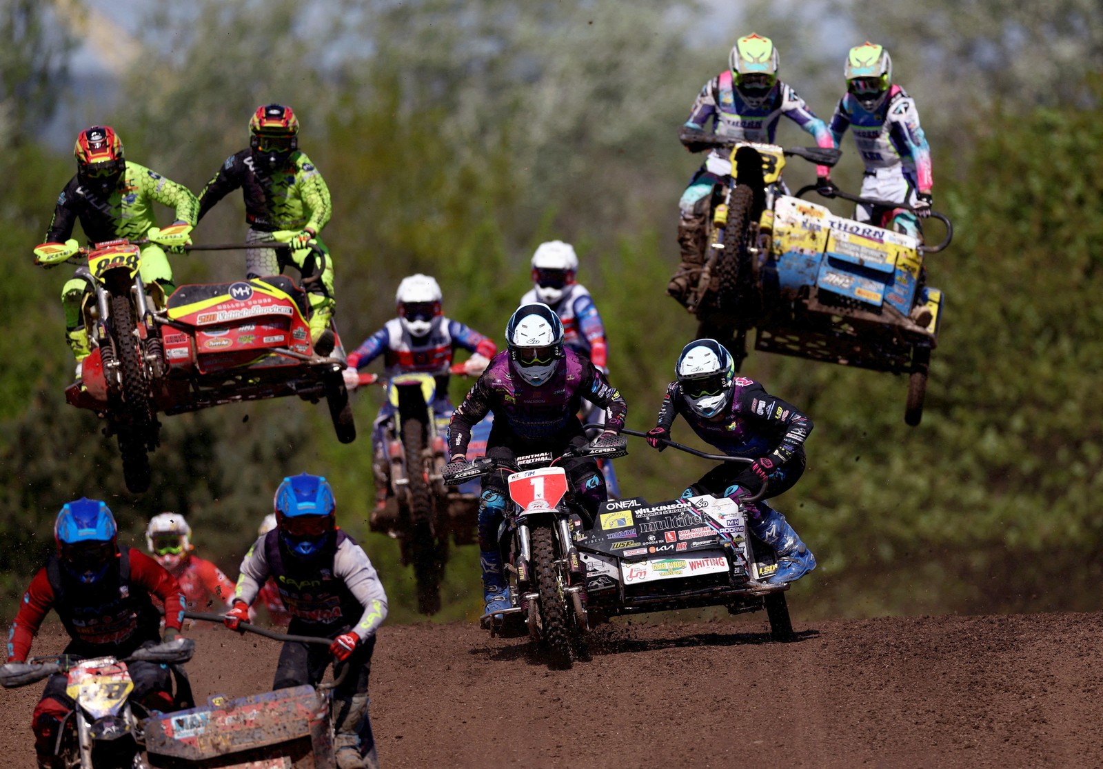 Sidecar motorcycle racers compete on a dirt track, with several seen in mid-air.