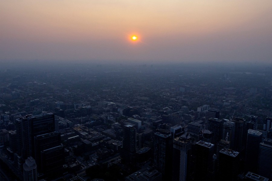 A reddish-colored sun is seen low in the sky, through hazy air above city streets.