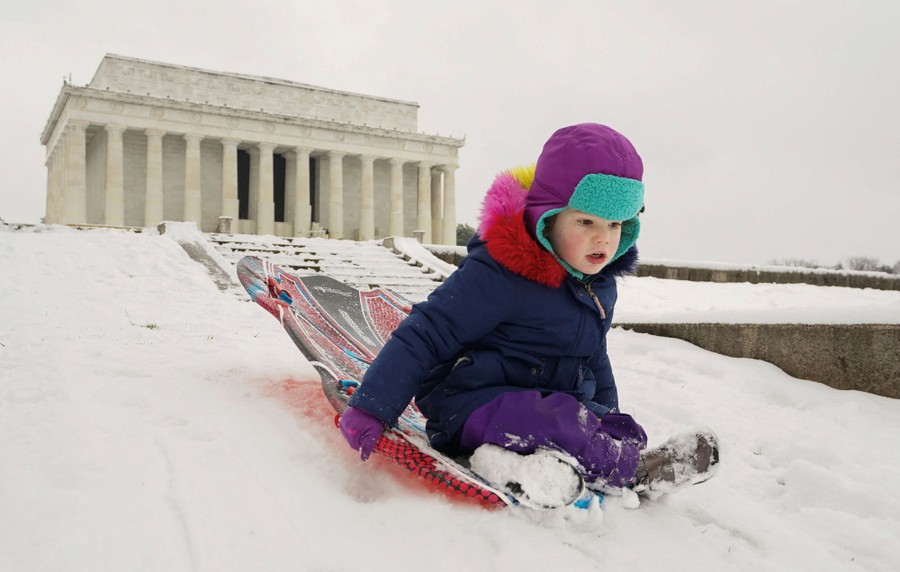 A child sleds down a snow-covered slope in front of the Lincoln Memorial.