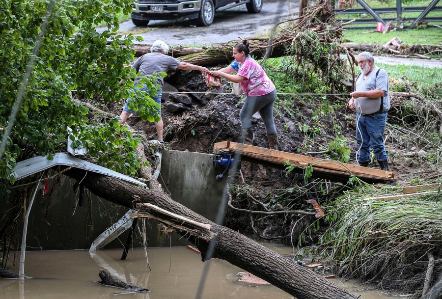 Three people work together on an embankment above a creek to transport food through flood debris.