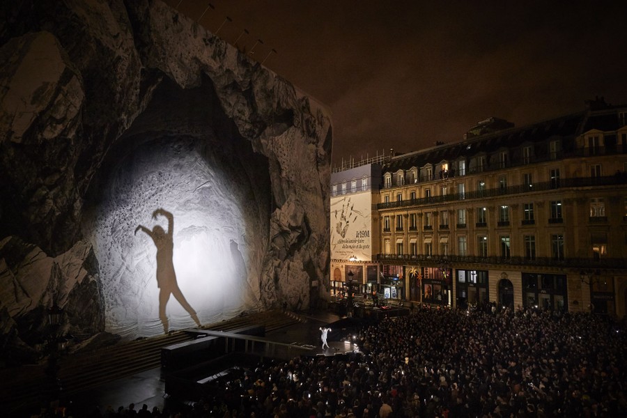 A dancer stands on a stage in front of a crowd in a city square. Bright light casts their huge shadow onto a decorated building behind them.