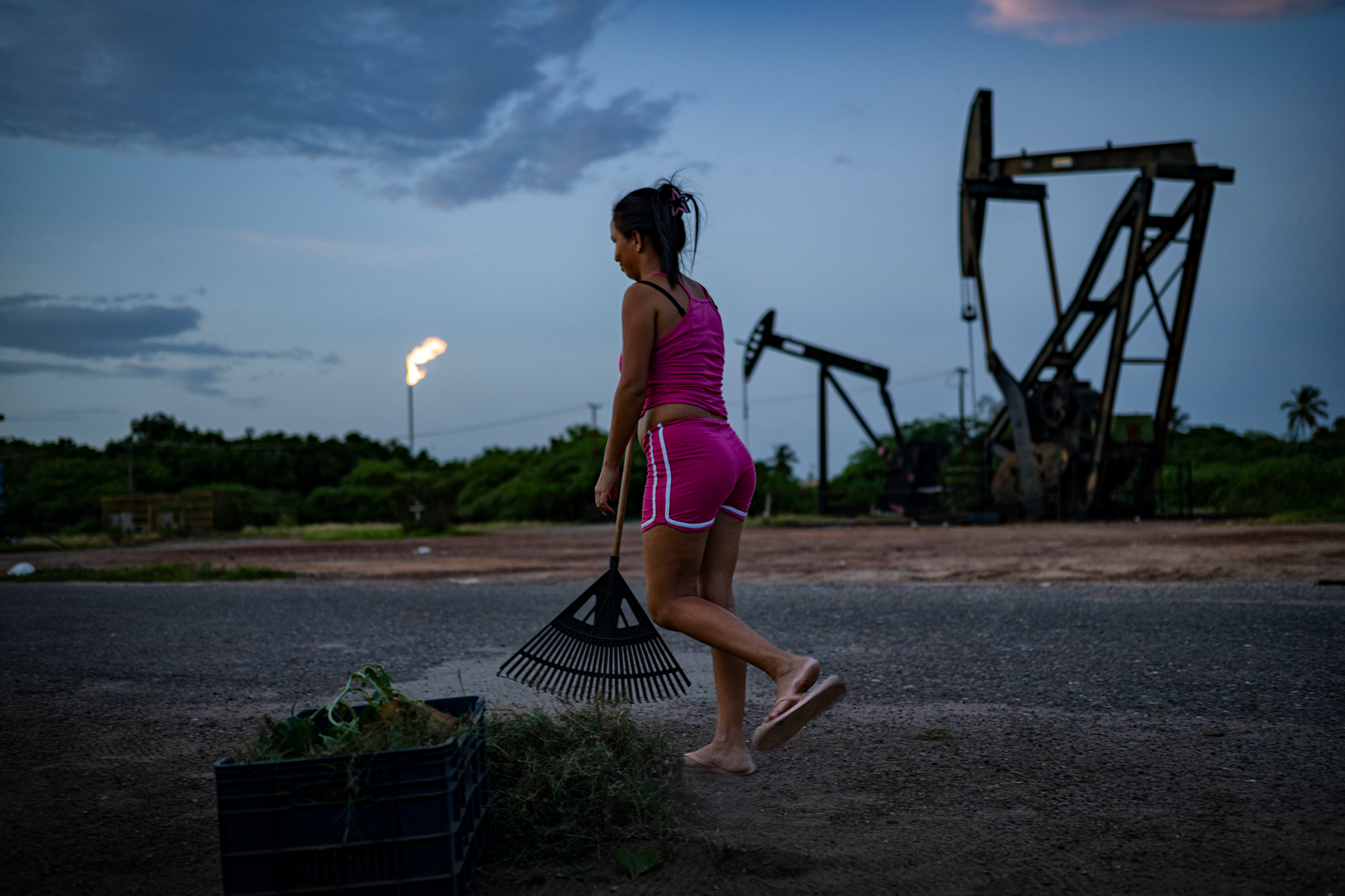 a woman wearing a pink shirt and orange shirt sweeps debris with a rake, with oil derricks in the background