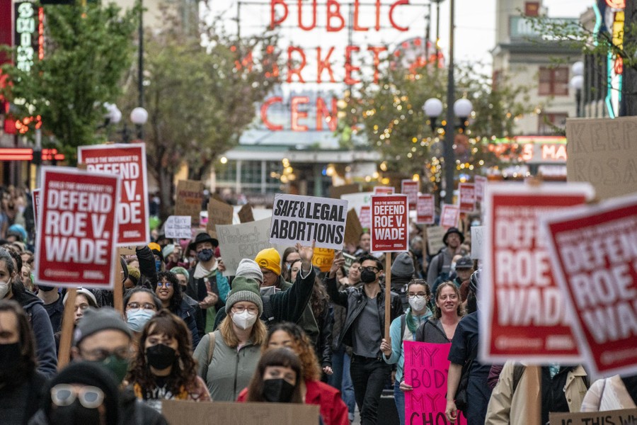 A crowd of protesters marches with signs in front of Seattle's Pike Place Market sign.