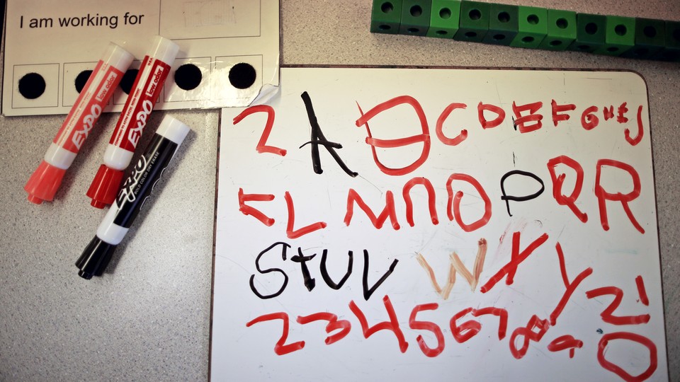 A whiteboard rests next to dry-erase markers and is covered in a student's attempts to scribble the alphabet.