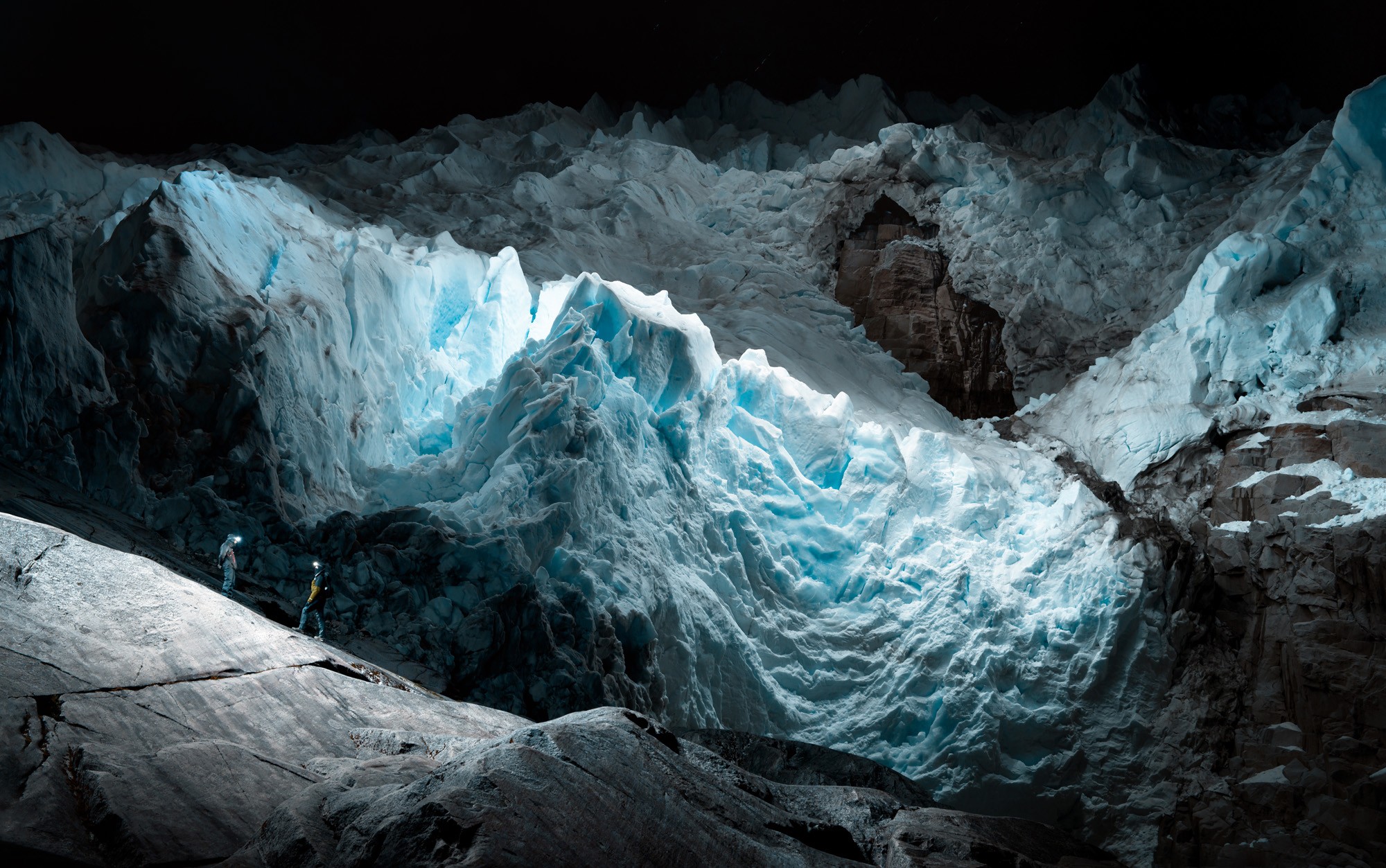 Two people stand on a smooth rock beside the craggy face of a glacier.