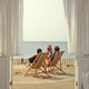 A hazy photograph of two people in beach chairs looking onto the ocean, taken through open patio doors from inside a house