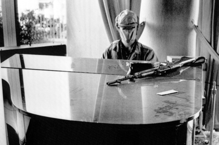 A masked person plays a piano, with a rifle resting nearby, atop the piano.