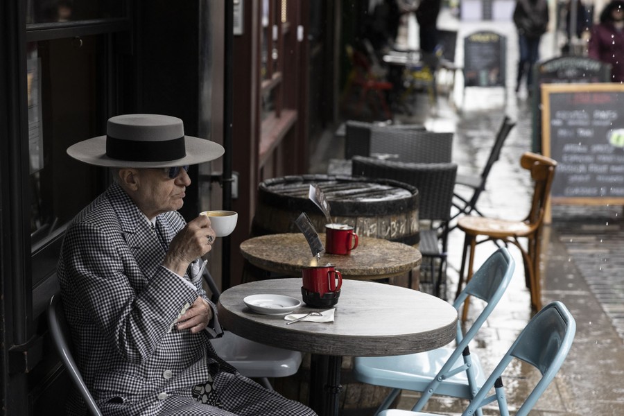 A stylishly dressed man sits at an outdoor café table, drinking coffee.