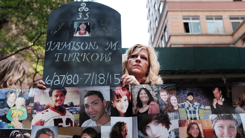 A woman holds a sign made to look like a gravestone next to photographs of young people.