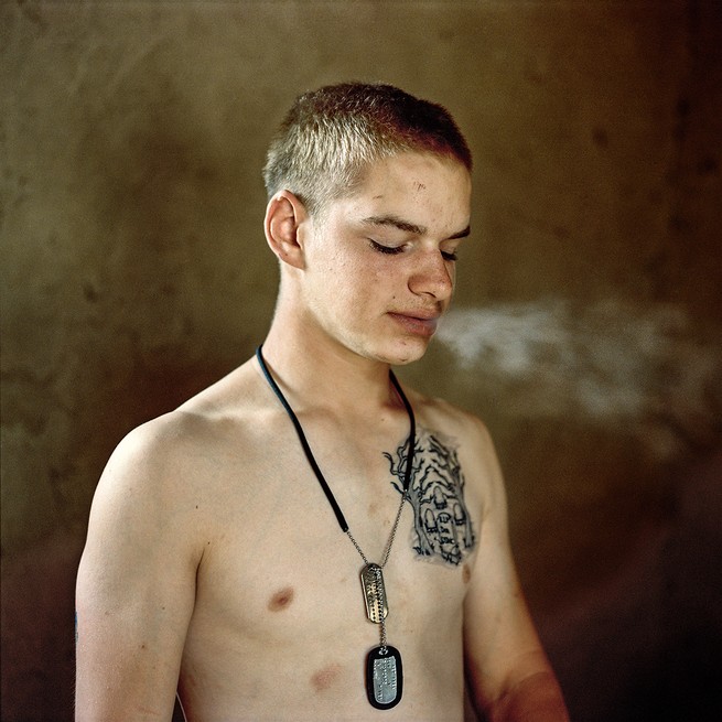 Shirtless young soldier with dogtags and large tattoo exhales cigarette smoke, looking down