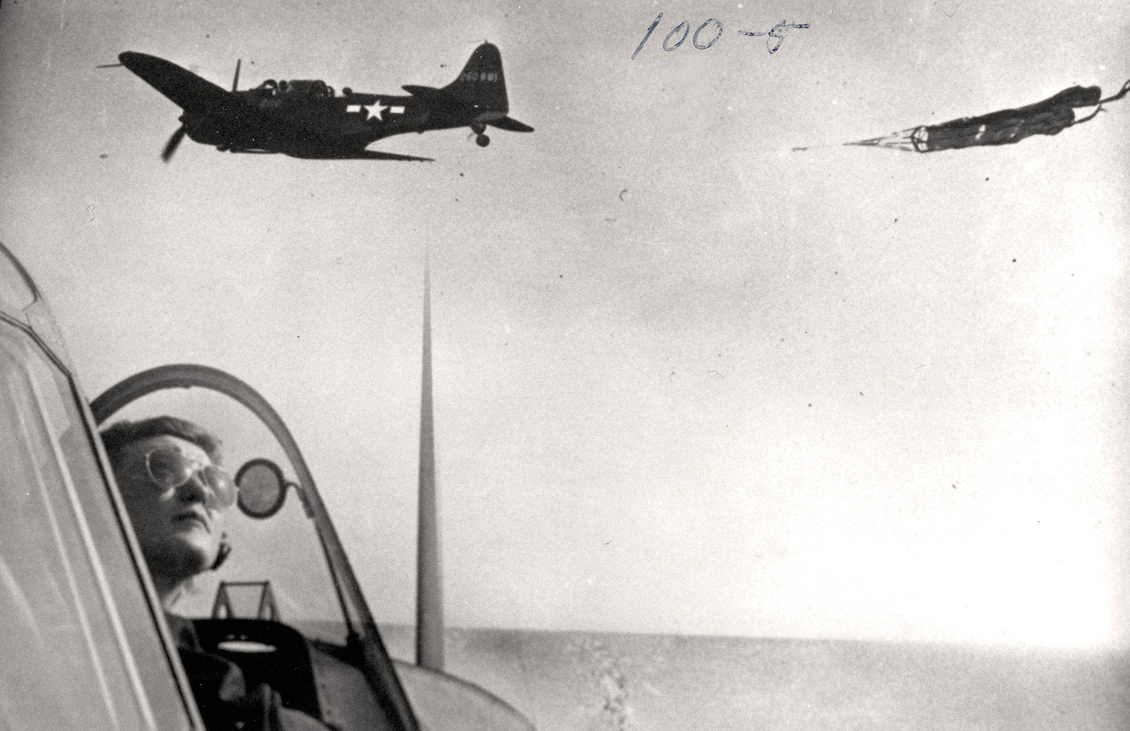 black-and-white archival photo of woman wearing aviators in open cockpit looking up as another plane towing a target passes perpendicularly overhead