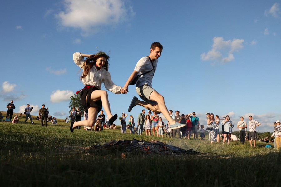 A couple jumps over a smoldering bonfire in a field.