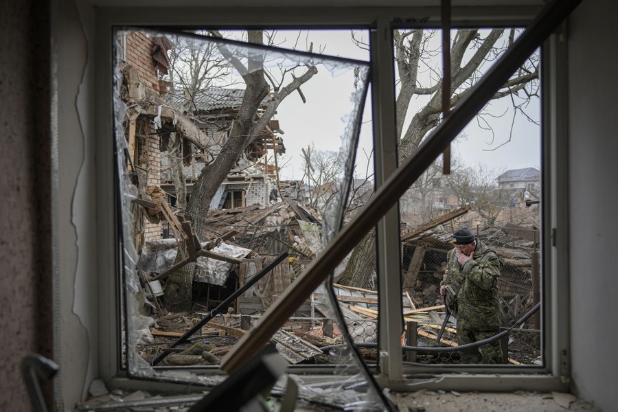 A shattered window frames a view outside of heavily-damaged houses and trees.