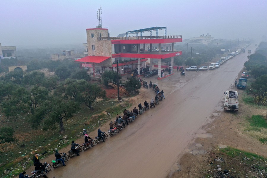 A long line of people with motorcycles stands outside a filling station.