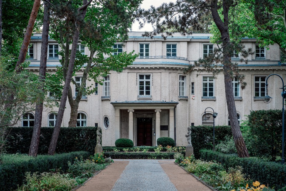 photo of large beige villa with many windows, columns by entrance, and trees flanking front walk 