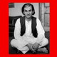 In a black-and-white image from 1988, Salman Rushdie, with chin-length dark hair and wearing a dark vest over white shirt and pants, sits crossed-legged on the floor.
