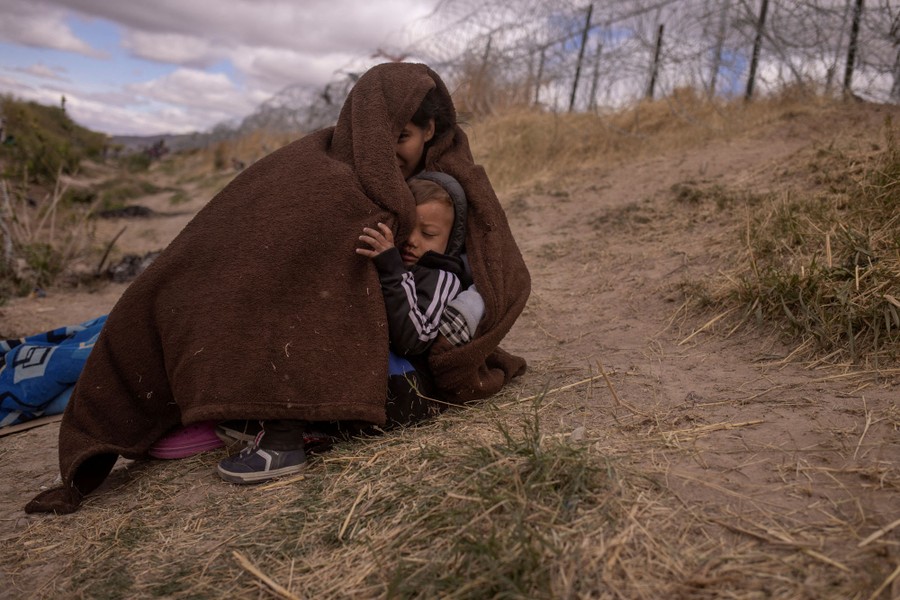 A young mother holds her child, both wrapped in a blanket, sitting on the ground near a border fence.
