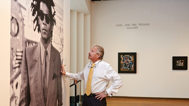 A man looking at a large portrait of Basquiat in museum gallery with two paintings hanging in the background.