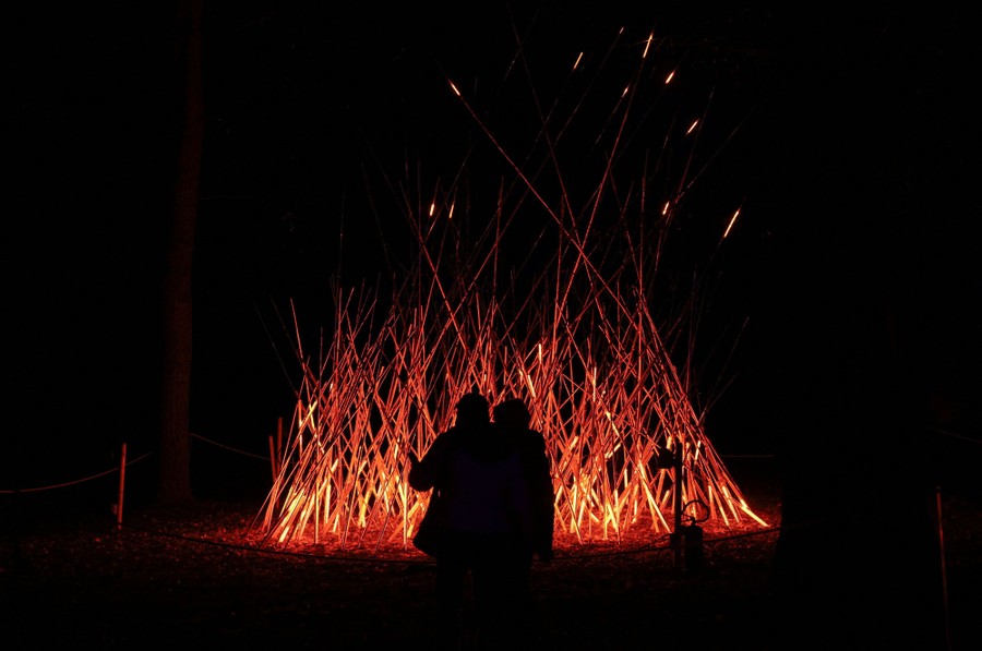 People stand near an art installation made of illuminated sticks, resembling a bonfire.