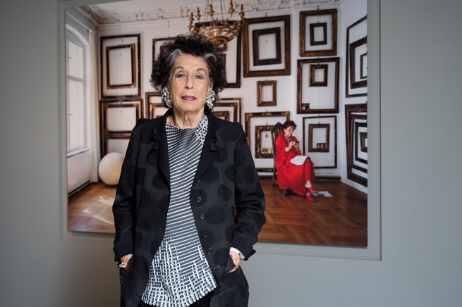 photo of woman in polka-dot black jacket stands with hands in pockets in front of a large photograph of herself seated, wearing red, in a large room full of empty wooden picture frames