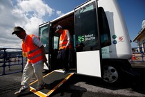 People in orange vests use a ramp to exit a driverless bus labeled "Robot Shuttle."